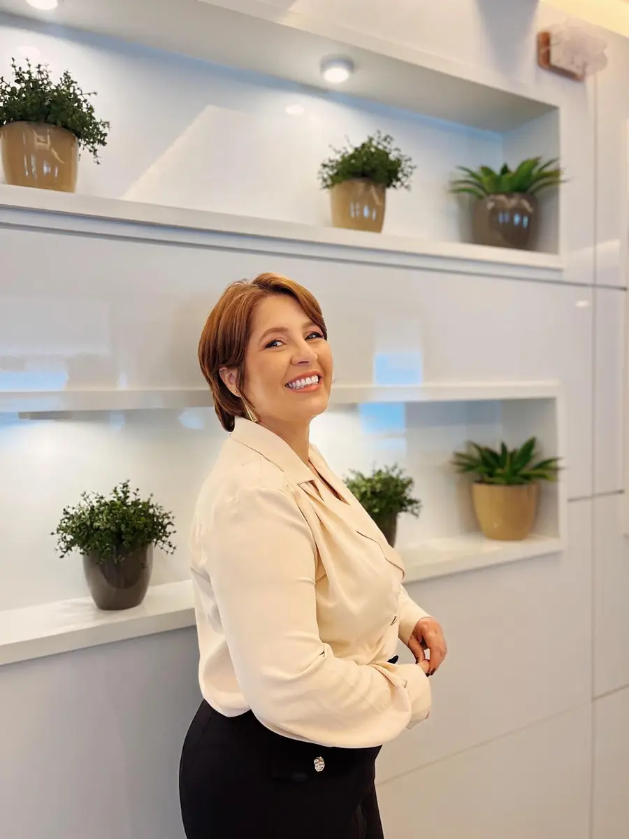 Ana Hercília em pé no consultório claro, sorrindo por sobre o ombro, com nichos brancos e plantas ao fundo