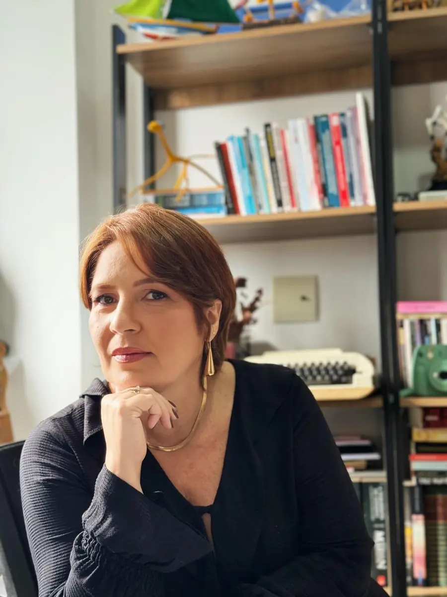 Ana Hercília com a mão no queixo e expressão atenta, diante de uma estante de livros